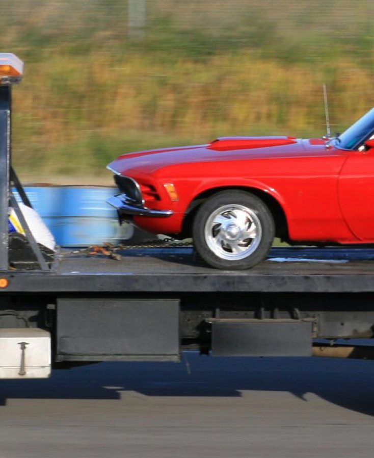 A red sports car being towed by a tow truck. Hauling, freight, or trucking theme. This image is a pan shot. Flat deck truck with automobile. Rural highway. White heavy duty truck with winch and loader. Flatbed.